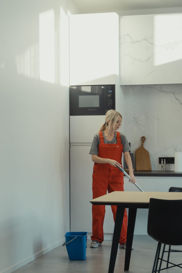 who-we-are A woman in red overalls mopping a kitchen floor under bright daylight, depicting housekeeping chores.